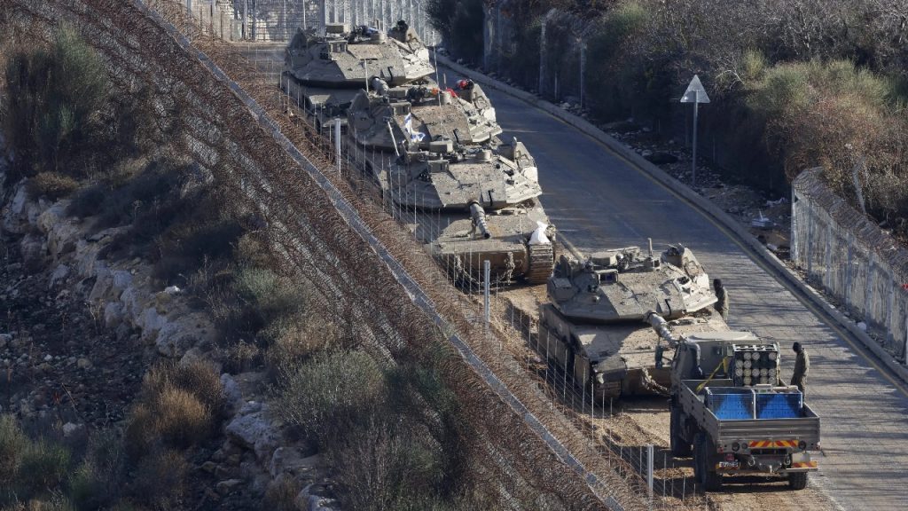 A line of Israeli Merkava tanks and a supply truck moving along a paved road next to a barbed-wire border fence