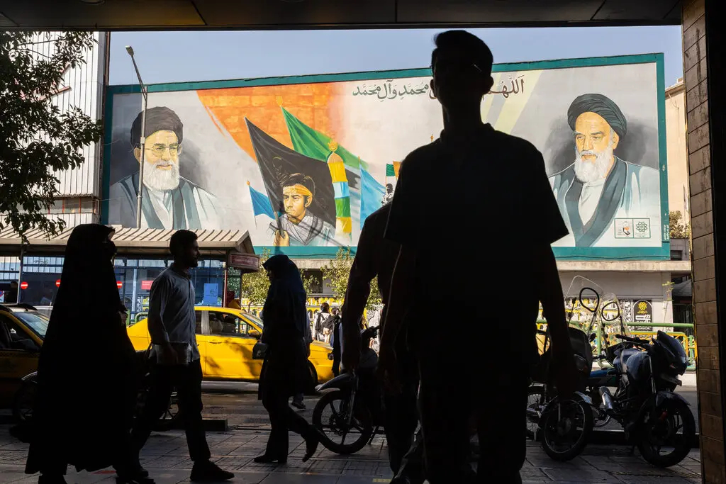 A man walking on a Tehran sidewalk with a large mural of Iranian leaders and a yellow taxi in the background