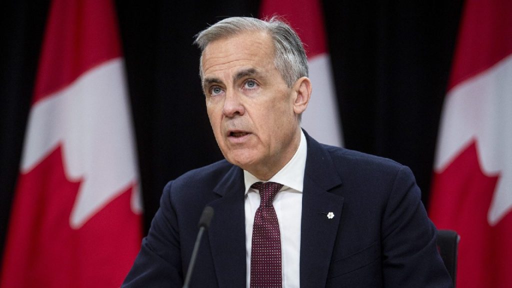 A man in a dark suit and red tie speaking at a podium with Canadian flags in the background.