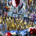 A dense memorial field filled with hundreds of small yellow and blue Ukrainian flags, interspersed with framed portraits of soldiers and colorful funeral flowers.