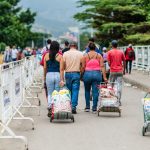 A medium shot from behind of people walking down a paved path, some pulling small metal carts filled with supplies, with white metal barriers on the left and lush green trees in the background.