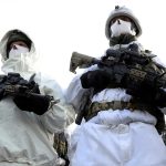 Two soldiers in full white winter camouflage gear, including white face masks and helmets, standing side-by-side and holding black assault rifles equipped with advanced scopes and tactical attachments.