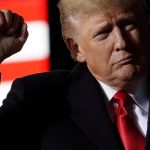 A close-up portrait of Donald Trump raising a clenched fist, wearing a dark coat and a bright red tie, with a blurred American flag featuring red and white stripes in the background.