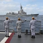 Five sailors in gray and white camouflage uniforms standing on the deck of a ship, facing away from the camera and saluting a large gray guided-missile destroyer with the hull number "41" sailing parallel to them in the open sea.