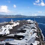 A high-angle view of a United States aircraft carrier deck at sea, packed with various fighter jets including F/A-18 Super Hornets, with a mountainous coastline and blue sky in the background.