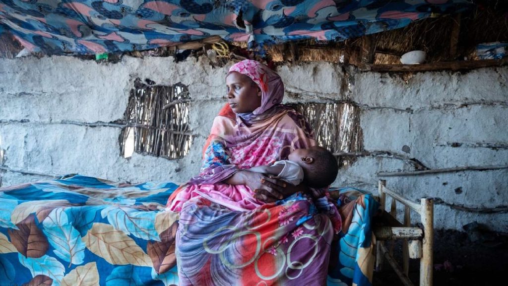 A woman in a colorful hijab holding a sleeping child inside a rustic, mud-walled shelter.
