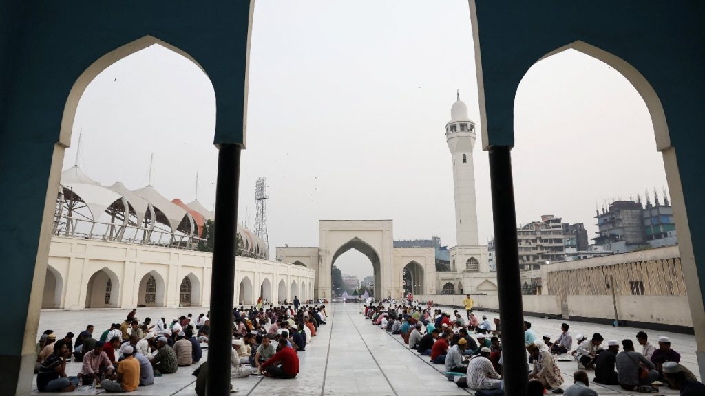 A wide view of many people sitting in rows on the ground in the courtyard of a large, white mosque with a tall minaret.