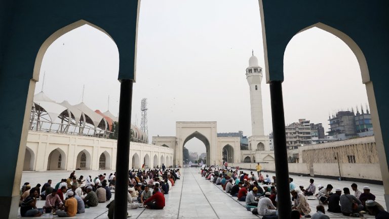 A wide view of many people sitting in rows on the ground in the courtyard of a large, white mosque with a tall minaret.