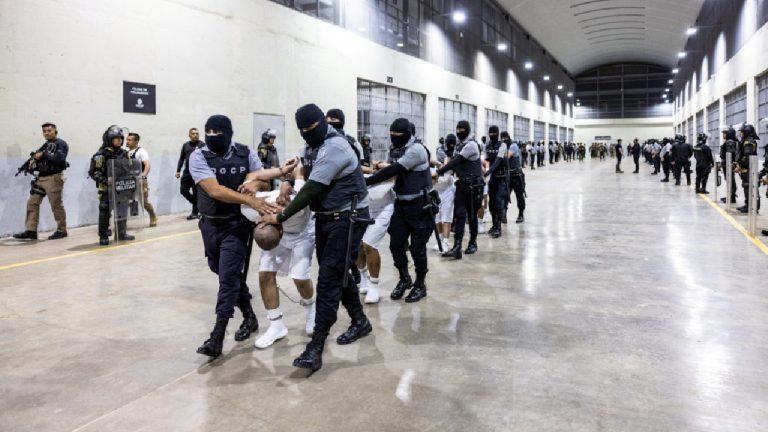 Masked security officers in El Salvador escorting a line of inmates in white uniforms through a large, high-security prison corridor.