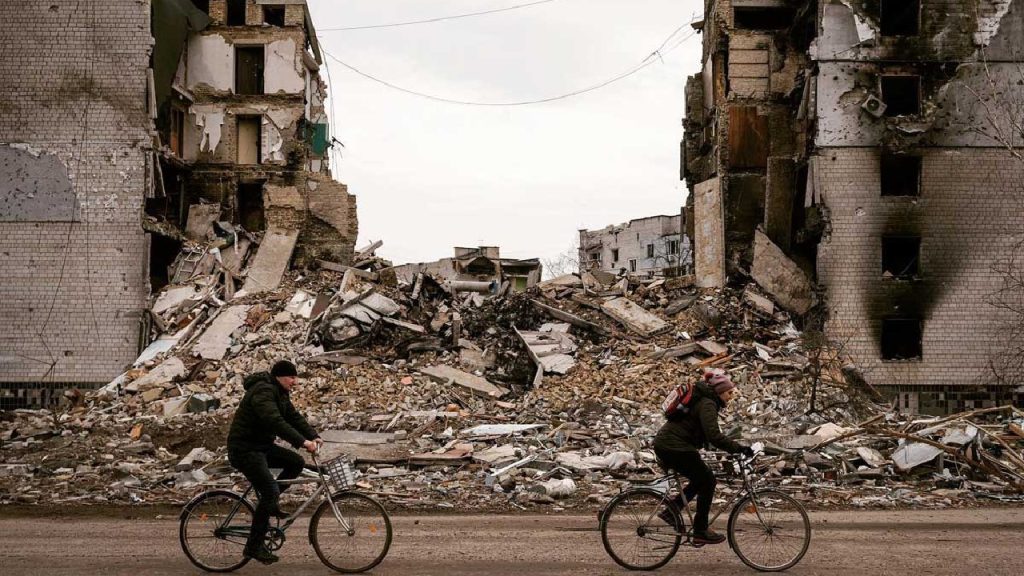 Two people riding bicycles past a heavily destroyed apartment building in a war zone.