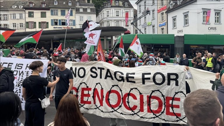 Protesters holding a large banner that reads "NO STAGE FOR GENOCIDE" with Palestinian flags in a crowded city square.