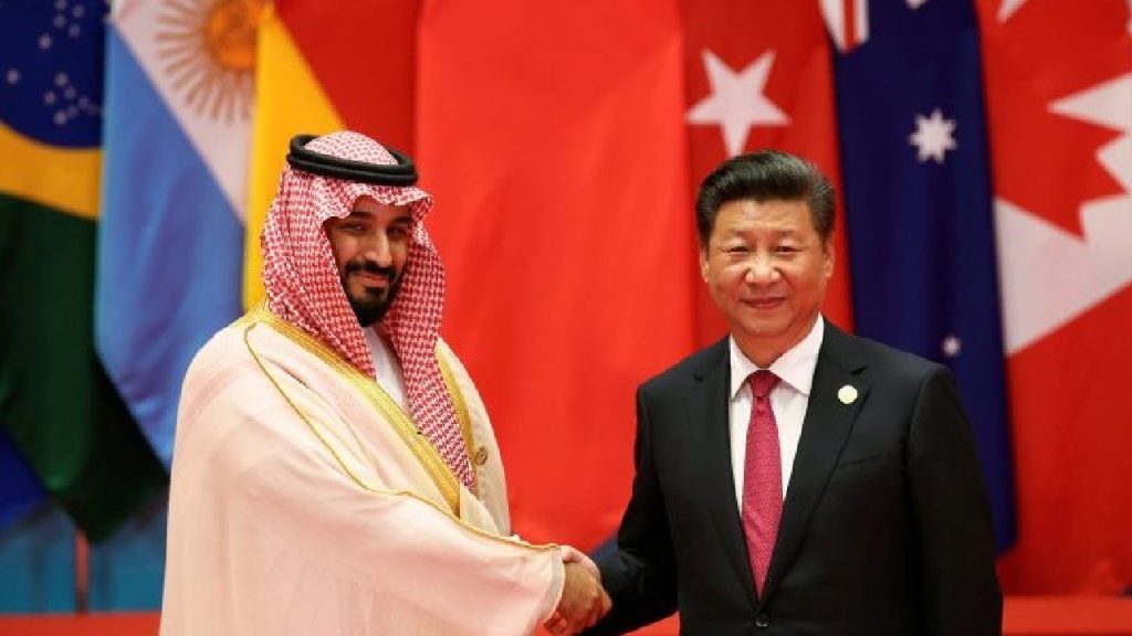 Mohammed bin Salman in traditional Saudi attire shaking hands with Xi Jinping in a black suit, standing in front of a row of international flags.