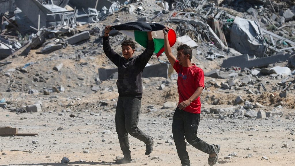 Two young men running through a field of gray rubble and destroyed buildings, smiling while holding up a Palestinian flag.