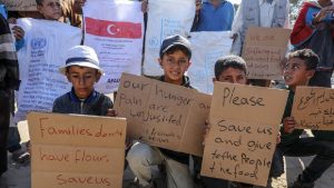 Children in Gaza holding handwritten cardboard signs appealing for food and help, with bags of aid in the background