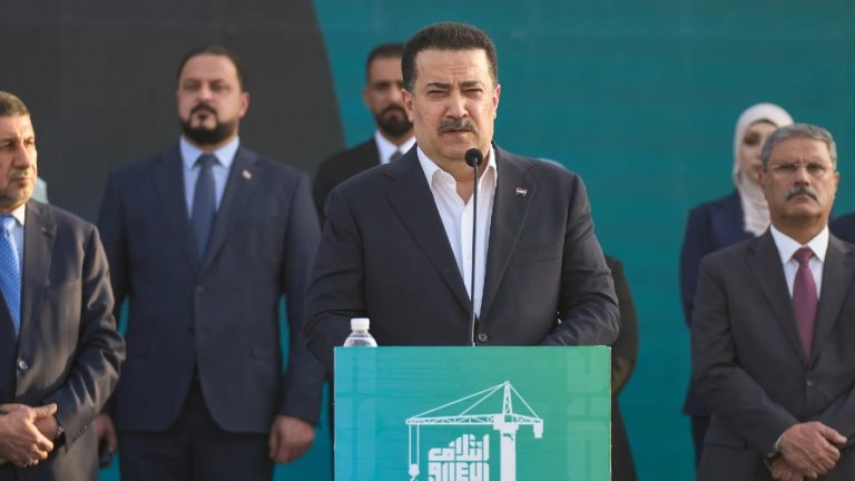 Iraqi Prime Minister Mohammed Shia' al-Sudani speaking at a green podium with a construction crane logo, surrounded by officials.