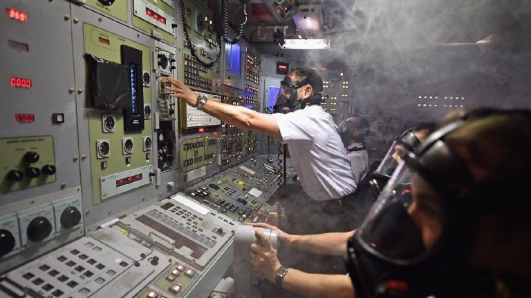 Several crew members wearing gas masks operating a complex control panel filled with buttons, dials, and digital displays in a smoky, dimly lit room.
