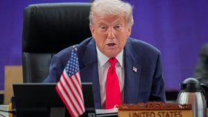 A close-up of Donald Trump speaking at a summit table with a "United States" nameplate and a small American flag in front of him