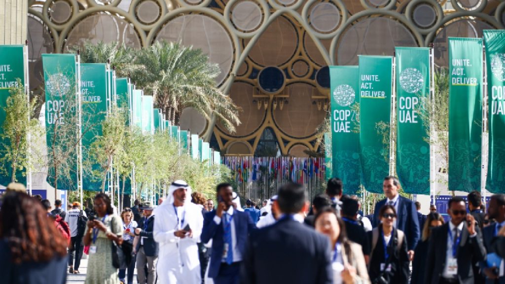 A crowded outdoor walkway lined with tall green "COP28 UAE" banners, with palm trees and a large domed architectural structure in the background.