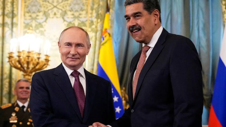 Vladimir Putin and Nicolas Maduro smiling and shaking hands in a formal room with flags.