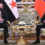 Vladimir Putin seated across a table from a Syrian official, in a formal meeting setting with flags in the background