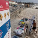 A wide shot of Bondi Beach in Sydney, Australia, featuring a safety warning sign in the foreground and a surfer walking across the sand