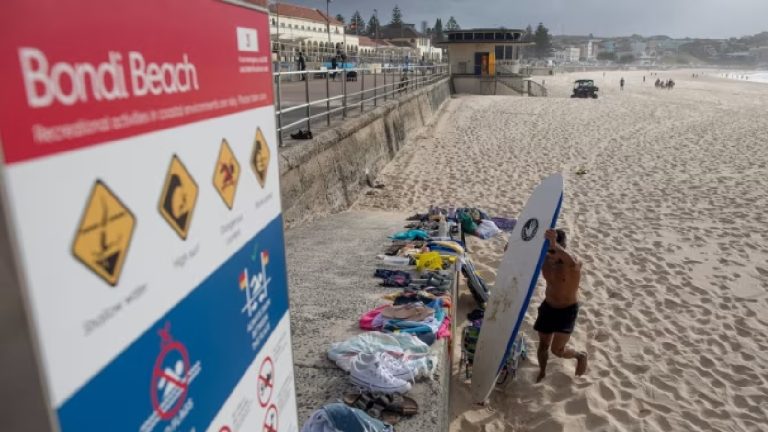 A wide shot of Bondi Beach in Sydney, Australia, featuring a safety warning sign in the foreground and a surfer walking across the sand