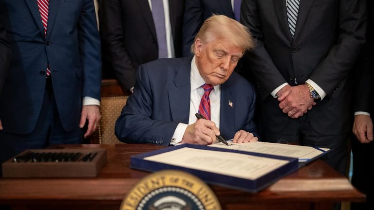 Donald Trump sitting at a dark wooden desk, focused on signing a formal document with a black pen, surrounded by people in suits.