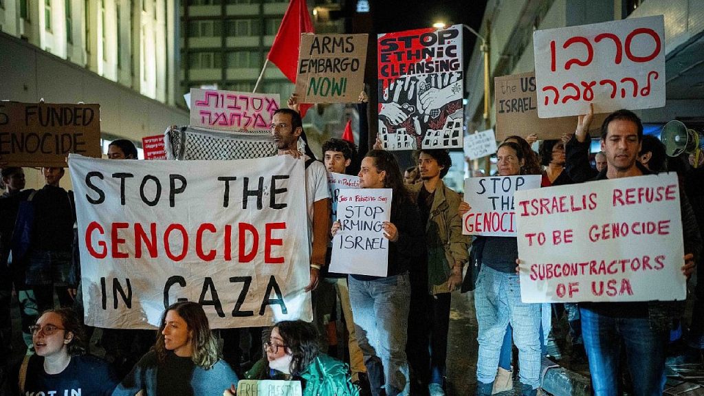 A large crowd of protesters at night holding signs in English and Hebrew.