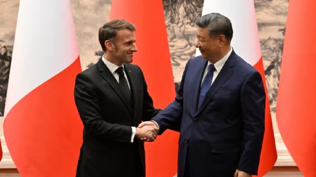 French President Emmanuel Macron and Chinese President Xi Jinping shaking hands and smiling warmly in front of national flags.