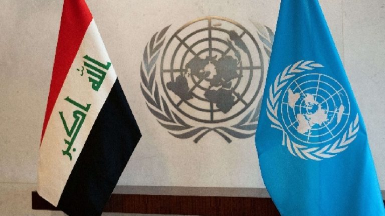 The flag of Iraq and the United Nations flag standing side-by-side in front of a wall with a large, faded UN emblem.