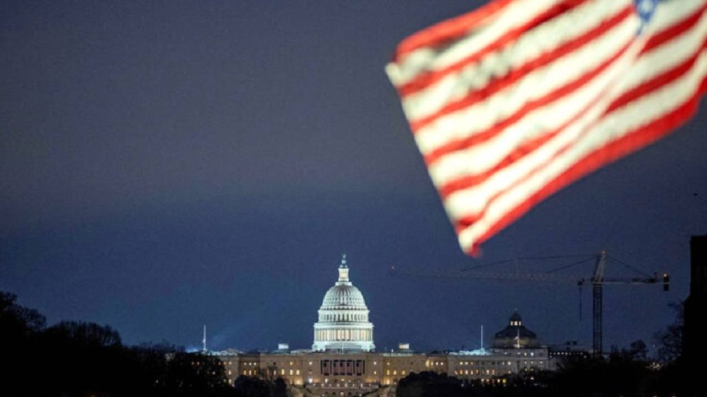 The US Capitol Building illuminated at night with a blurred American flag in the foreground.