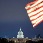 The US Capitol Building illuminated at night with a blurred American flag in the foreground.