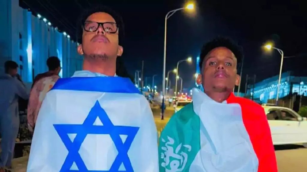 Two young men standing outdoors at night; one is wrapped in the flag of Israel and the other is wrapped in the flag of Somaliland.
