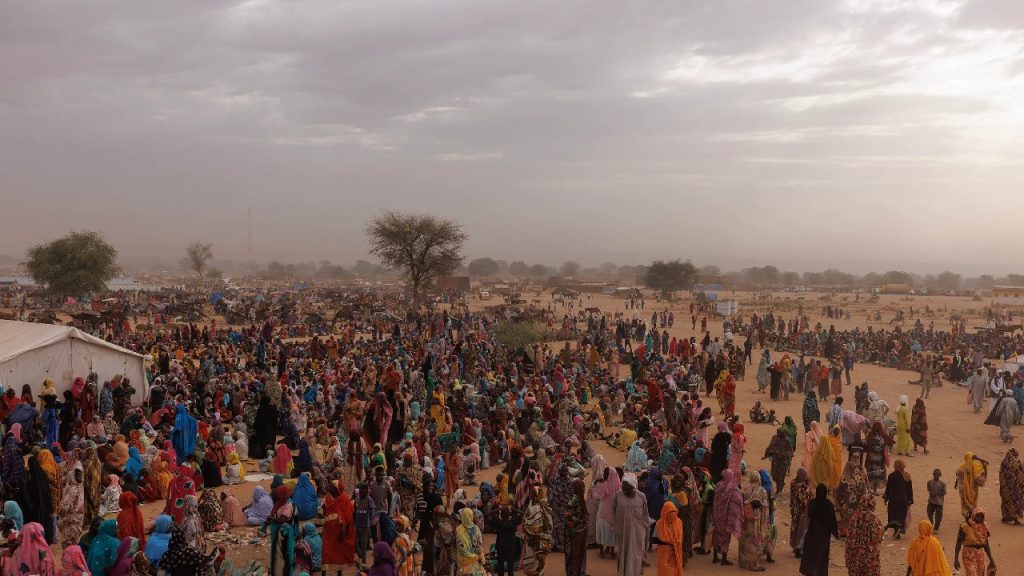 A vast, crowded displacement camp in Sudan under a hazy sky, showing thousands of people fleeing the ongoing genocide and humanitarian crisis.