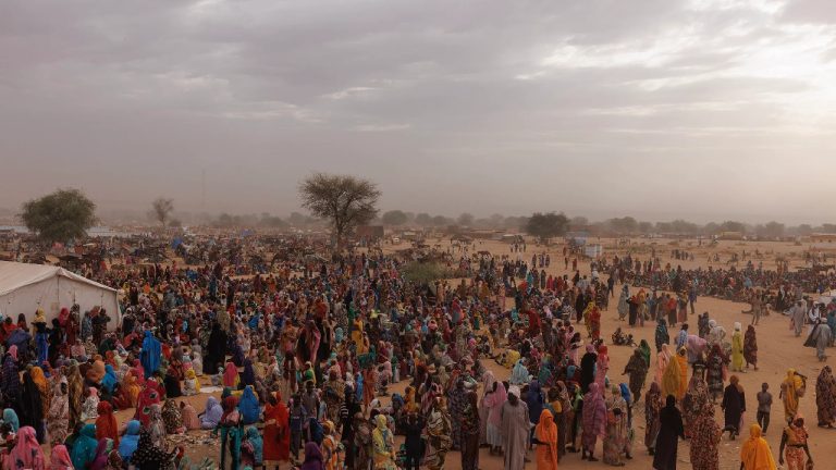 A vast, crowded displacement camp in Sudan under a hazy sky, showing thousands of people fleeing the ongoing genocide and humanitarian crisis.