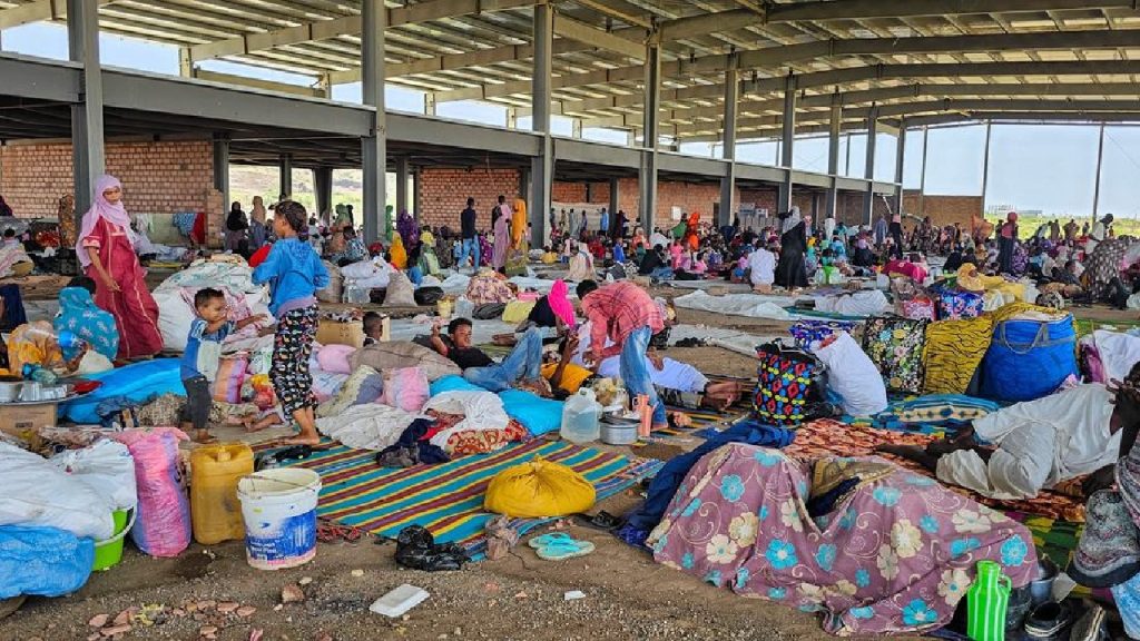 Hundreds of displaced Sudanese people taking shelter with their belongings in a large, open-air industrial structure.