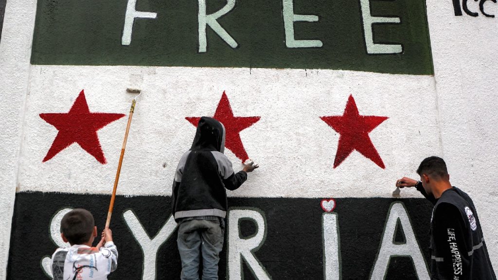 Three people, including two children, painting the "Free Syria" flag on a large white wall.