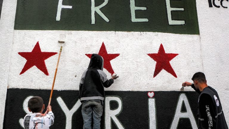 Three people, including two children, painting the "Free Syria" flag on a large white wall.