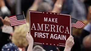 Hands holding a maroon sign that reads "MAKE AMERICA FIRST AGAIN" and two small American flags at a rally