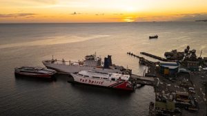 Sunset view of a busy port with cargo ships and ferries, including the "T&T Spirit," docked near an industrial area