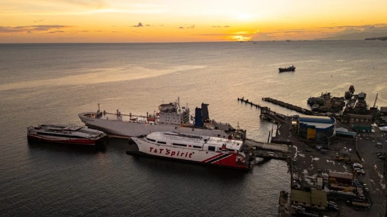 Sunset view of a busy port with cargo ships and ferries, including the "T&T Spirit," docked near an industrial area