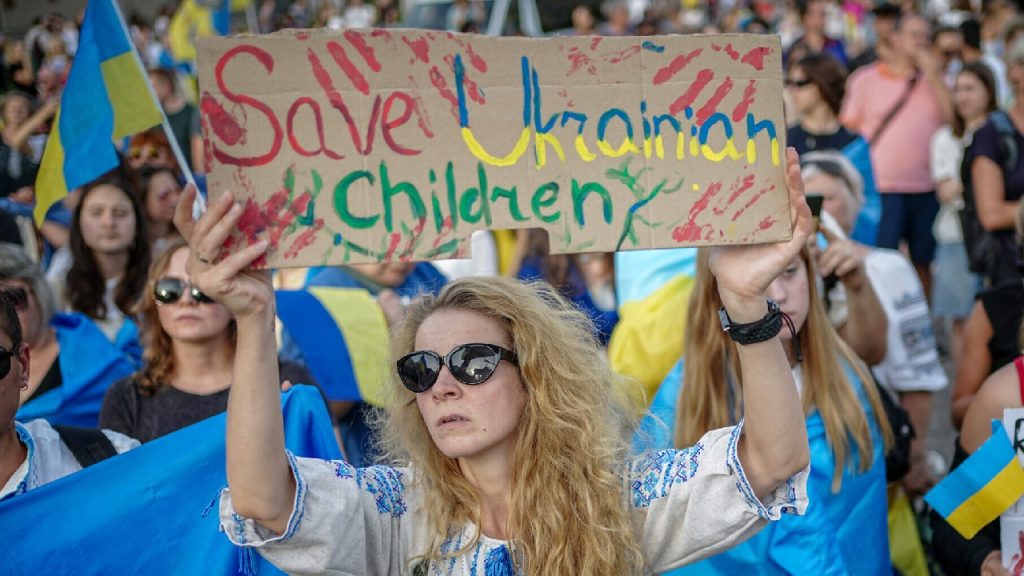 Woman wearing sunglasses and an embroidered shirt holding a sign that reads "Save Ukrainian Children" at a public protest