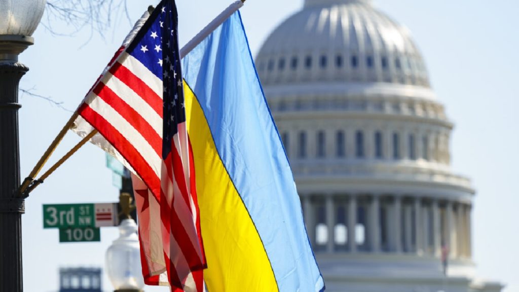 US and Ukrainian flags flying side-by-side on a pole, with the dome of the US Capitol Building blurred in the background
