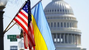 US and Ukrainian flags flying side-by-side on a pole, with the dome of the US Capitol Building blurred in the background