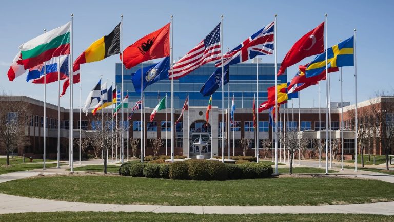 A wide shot of various national flags, including the US, UK, and Turkey, flying in a circle in front of a modern brick and glass building.