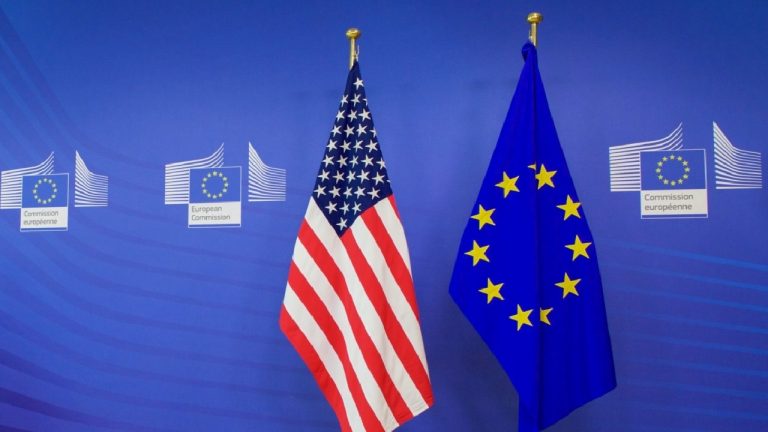 The United States flag and the European Union flag standing side-by-side on gold-topped poles against a blue wall featuring the European Commission logo.