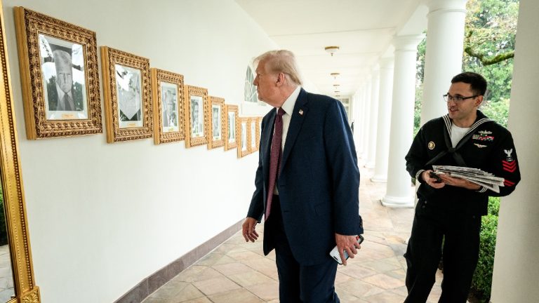 Donald Trump walking down a White House colonnade past framed portraits, followed by a military aide.