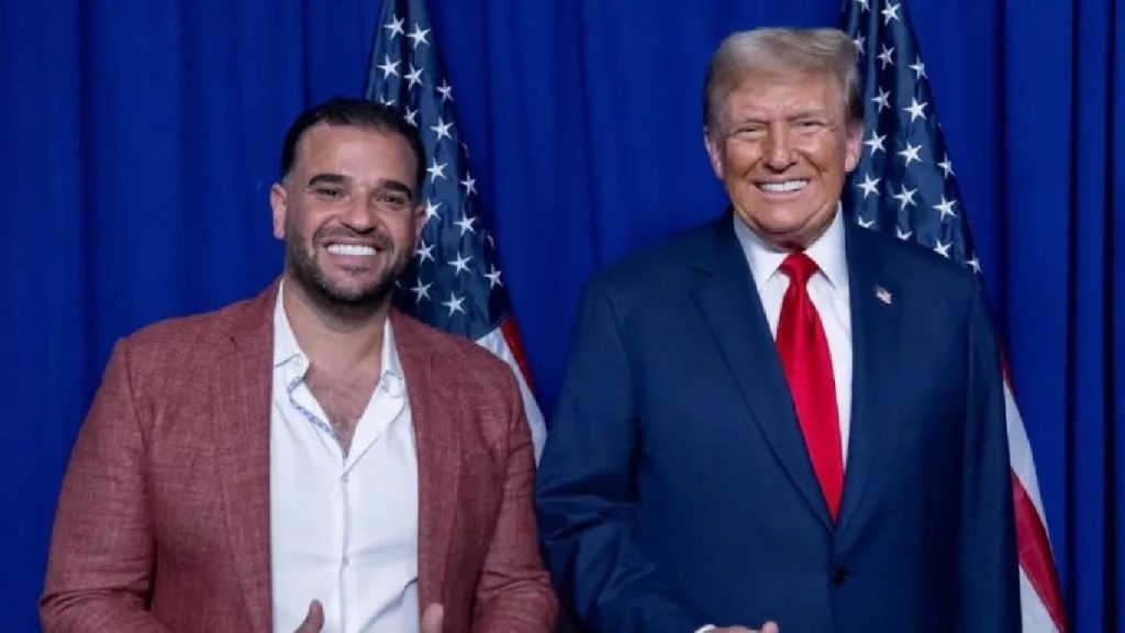 Donald Trump in a navy suit and red tie standing next to a smiling man in a reddish-brown blazer in front of two American flags and a blue curtain background.