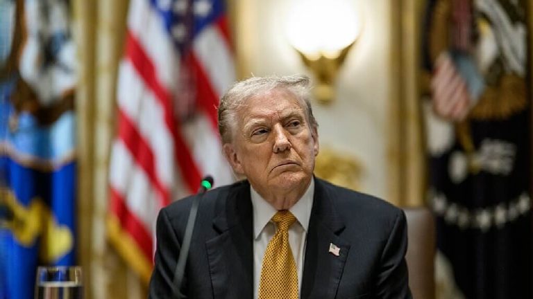 Donald Trump seated at a formal table, looking serious, with American flags visible in the background