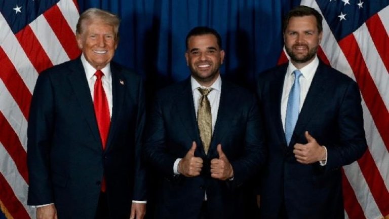 Donald Trump, Mark Savaya, and JD Vance standing together and smiling in front of American flags.